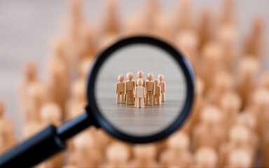 Fototapeta premium A magnifying glass is focusing on a small group of wooden figures standing out from a larger blurred group, highlighting themes of selection and individuality.