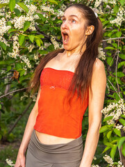 Young woman sneezes in the park against the background of a flowering tree. Allergy to pollen...