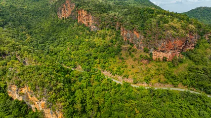 Poster Aerial panoramic view of lush green hills and valleys intersected by a winding mountain road. The landscape features rugged cliffs, dense forests, and a distant horizon under a partly cloudy sky.