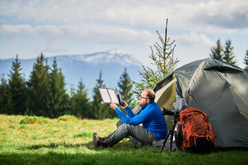 Man sits beside tourist tent, holding portable solar panel charger to capture sunlight for charging...