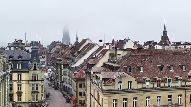 Foggy misty morning panorama overlooking the historic rooftops of Bern's Old Town, Switzerland, with the iconic Munster Cathedral (Berner Münster) spire rising through the atmospheric mist