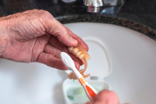 An elderly person scrubs a partial denture with a toothbrush over a bathroom sink, demonstrating proper denture cleaning and oral hygiene maintenance.