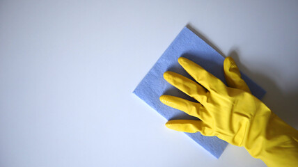 Photo of cleaning process. Hand in a rubber glove wiping a surface with a cellulose sponge. Yellow glove, blue lavender sponge. White surface. Copy space. Hand placed on the right side of background.
