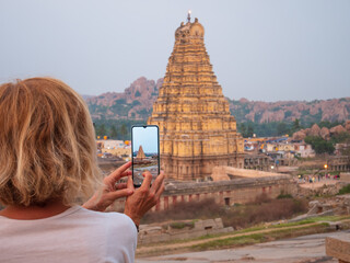Woman taking photo of amazing temples at Hampi, world famous unesco site. Tourist using mobile phone among ruins stone walls gateways in Karnataka, India