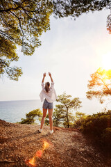 Woman enjoying sunset by the ocean with arms raised in the air, surrounded by trees and nature
