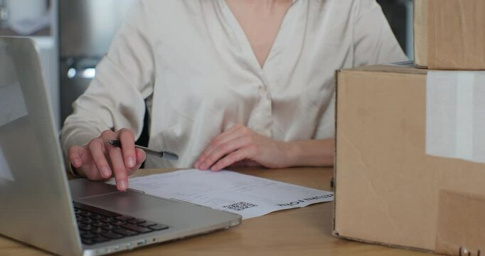 A young woman writes a return order and sits at a table with a laptop at home. Close-up video of a female customer writing on paper and filling out a form, wanting to return cardboard box back.