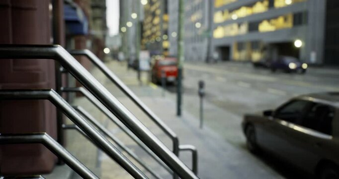 Steel handrails stand prominently in the foreground, leading to a quiet city street lined with parked cars. Dusk settles over the city, casting a soft glow on the buildings.