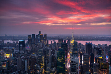 Fototapeta premium Aerial view of Manhattan at dusk with a tall illuminated skyscraper in the center. The city stretches toward the river under a soft pink and purple sunset sky.
