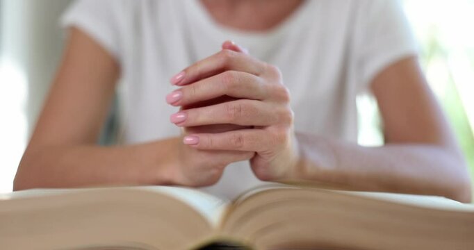 Worried woman prays holding hands over open thick old book in room. Lady with troubles seeks help in spiritual practices at home. Religion ritual
