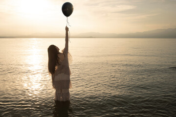 woman from behind holding a black balloon by the string who wants to escape over the infinite sea