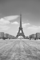 The Eiffel Tower rises majestically over a lush green park, surrounded by meticulously shaped hedges. Visitors stroll leisurely beneath the iconic structure on a sunny day.