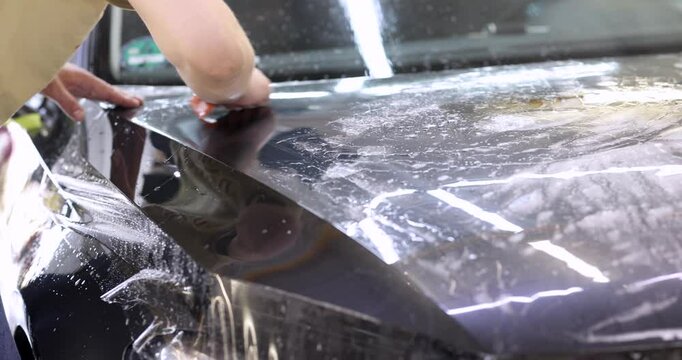 Man aligns section of chrome wrap with surface of car bonnet. Male begins smoothing chrome film carefully with plastic squeegee. Slow motion