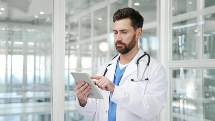 Confident doctor in white coat using digital tablet while standing at workplace in a modern hospital office. Focused handsome medical worker physician chats online with patient or works in application - Powered by Adobe