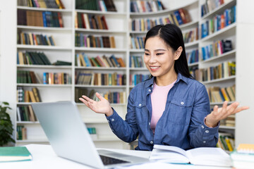 A smiling asian woman sits at a desk in a library, gesturing while looking at her laptop....
