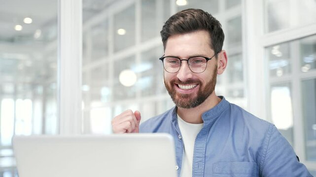 Satisfied happy businessman received great news on laptop while sitting at workplace in modern business office. Smiling glad handsome entrepreneur reads a positive good message on a computer. Close up