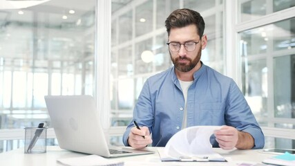 Confident businessman signing documents sitting at a desk at workplace in a business office. Executive in glasses looks through the folder with documentation, checks and signs the contract with a pen