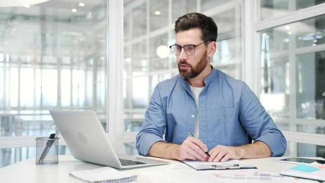 Busy businessman doing paperwork using laptop sitting at desk at workplace in business office. Confident financier working fills out documents, writing, making a tax return or engaged in accounting