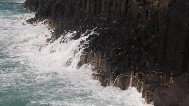 Swell surges, followed by larger waves, crash into the basalt cliffs with their iconic hexagonal columns, then run off the rocks at Fingal Head, a popular tourist spot in New South Wales, Australia.