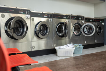 Row of washing machines in public laundromat and basket with dirty clothing on the floor