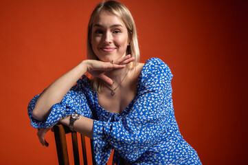 Confident, emotional young girl in blue floral dress leaning on chair on orange background....