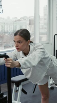 Vertical shot of brunette girl doing kickbacks on cable machine while recovering from illness or injury in rehabilitation facility