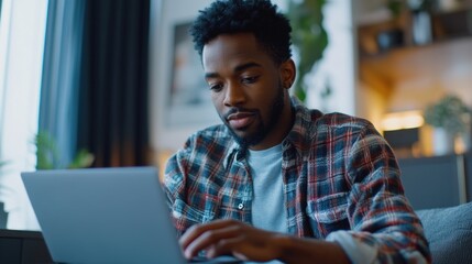 A man is sitting on a couch and using a laptop. He is wearing a plaid shirt and has a beard. Concept of relaxation and leisure, as the man is using his laptop in a comfortable setting