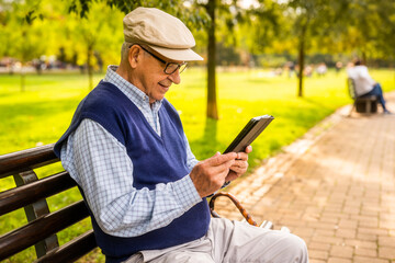 Obraz premium Old man with hat is sitting on bench in park on sunny day and using digital tablet.