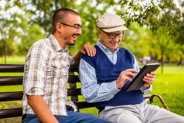 Obraz premium Portrait of happy grandfather and grandson in park. They are spending time together and using digital tablet.