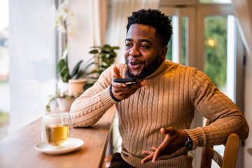 A young black man sits at the bar, casually dressed. He is using smartphone and recording voice...