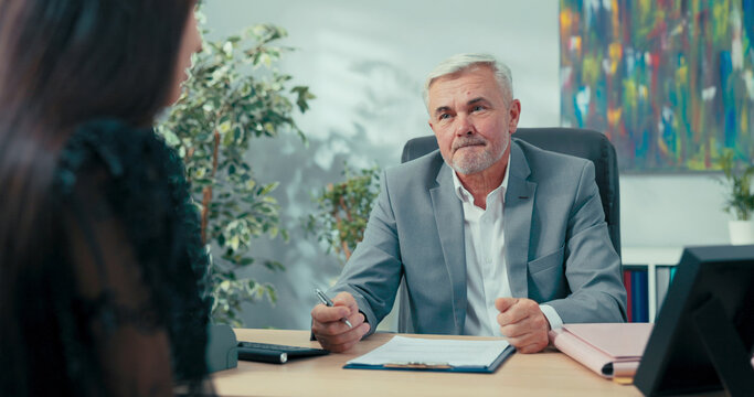 Job interview elderly boss with gray hair sitting at desk opposite him woman applying for secretarial position in company man asks girl questions discusses education salary expectations first contract