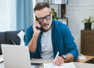 Obraz premium Man, phonecall and home office writing in notebook, mature and business person or taking notes with virtual contact. Schedule appointment, online and conversation with laptop, communication and talk