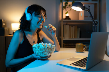 Caucasian woman watching movie eating popcorn, laughing, using laptop at home.