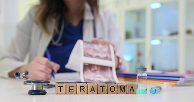 Wooden blocks with word Teratoma on desk and woman in coat and stethoscope holding ultrasound images. Medical context related to pathology