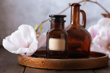 A dark bottle for herbalist oil or serum with Magnolia on a dark brown plate on grey light background. Herbalist theme. Alternative medicine. Healthy plant. 