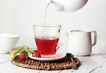 Female hand holding a teapot and pouring tea into a transparent glass cup with a raspberries. On a white plate on a wooden background.