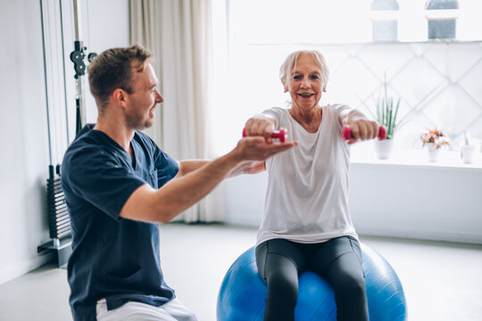 Senior woman lifting weights with physiotherapist on fitness ball: rehabilitation and healthy lifestyle