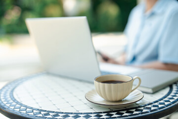 Relaxing outdoor workspace with coffee cup on foreground and blurred casual woman using laptop on background. Modern remote working, comfortable leisure and digital lifestyle concept.