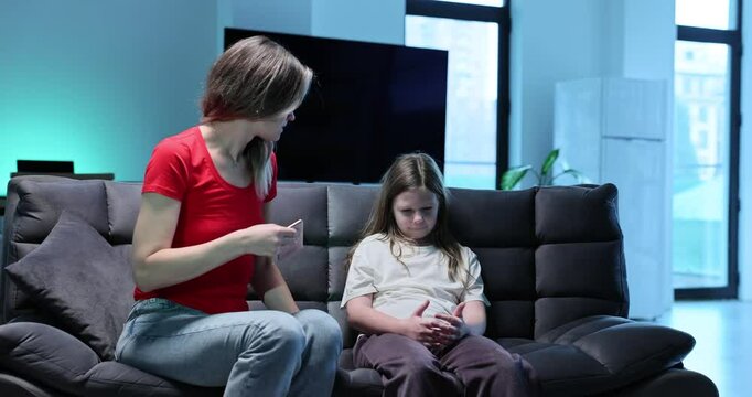 Angry mother scolds daughter and takes away smartphone in living room. Little girl addicted from gadget and emotional female parent sit on sofa