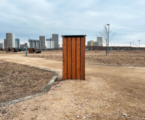 Wooden trash can in the foreground of an urban park, pathways and barren trees visible. High-rise...