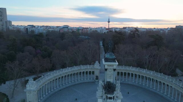 Breathtaking drone shot flying toward the statue at Retiro Park's pond during sunrise. The skyline, including the iconic Torrespa&ntilde;a tower, is visible in the background, with warm morning light