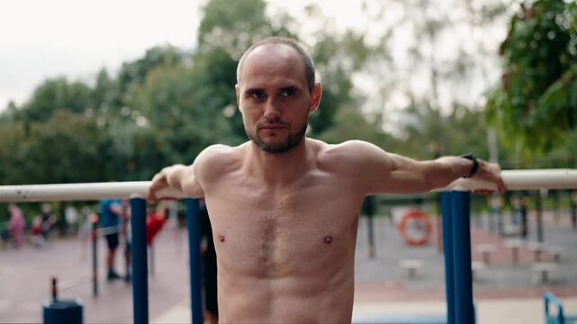 A shirtless man stretches his arms on parallel bars at an outdoor gym. He smiles as he breathes deeply and enjoys the fresh air and sunshine. Perfect for fitness, health, and wellness campaigns.