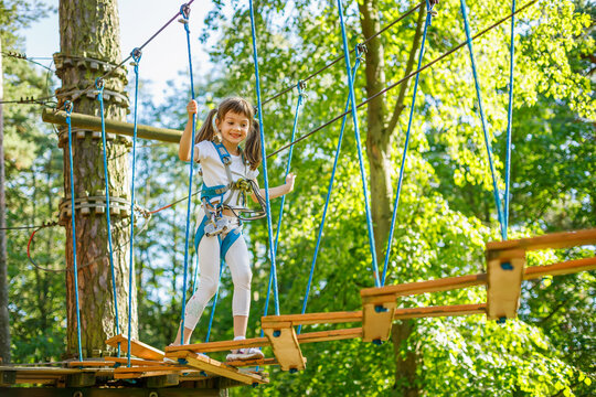 Young girl navigating a ropes course in an adventure park, smiling confidently while wearing a safety harness and balancing between platforms