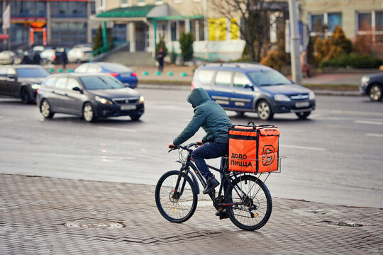 Minsk, Belarus. Dec 17, 2024. Bicycle food delivery courier riding through busy urban street with an insulated backpack, fast service. Dodo pizza delivery. Cyclist food courier braving rainy weather