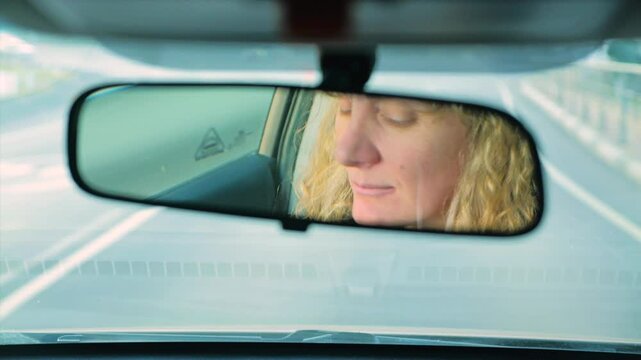 Close-up of a woman with long blonde hair driving a car while looking in the rearview mirror. A highway stretches ahead through the dirty windshield