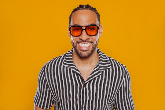 Studio portrait of a cheerful young multiracial man with braided hair, wearing sunglasses and a stylish striped shirt, posing against a vibrant yellow background.