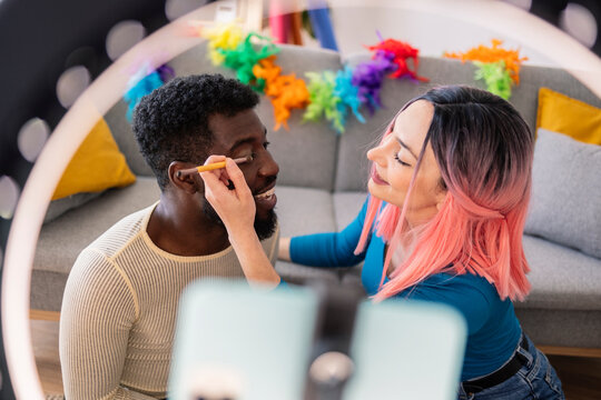 Woman doing eye-make up of non-binary friend seen through ring light