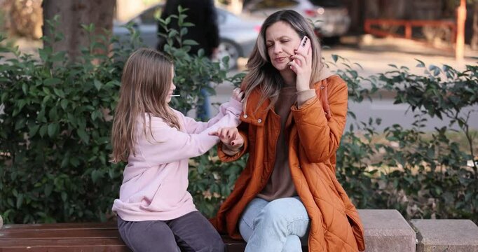 Woman holds phone to ear and engages in conversation sitting on bench. Little daughter persistently tugs at sleeve of mom to get attention slow motion