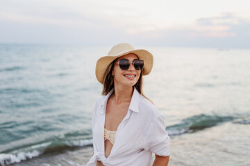 Smiling young woman at the beach wearing sunglasses and a straw hat while enjoying a sunset by the...