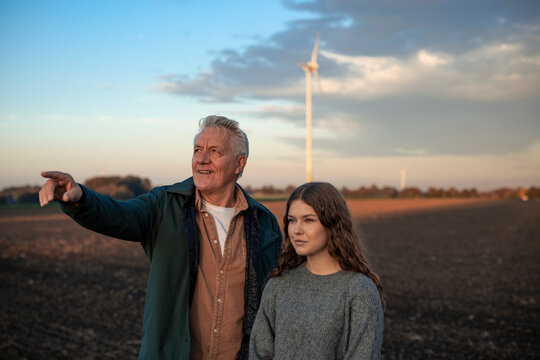 Grandfather and granddaughter in a field with a wind turbine at sunset