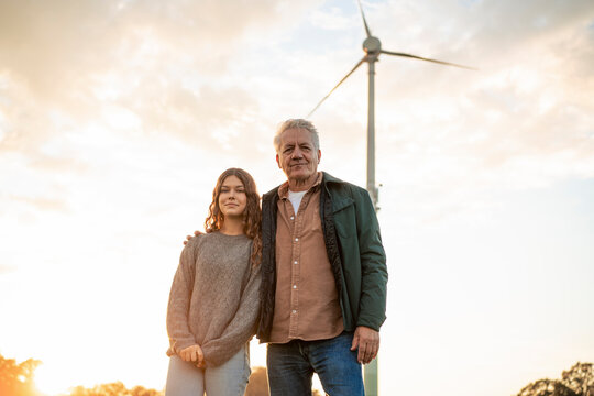 Grandfather and granddaughter standing together near a wind turbine at sunset